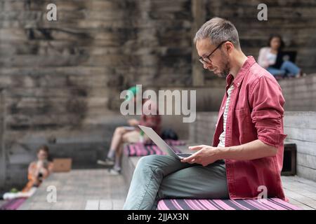 Young focused student guy with laptop sitting on wooden bench outside. Outdoor coworking space Stock Photo