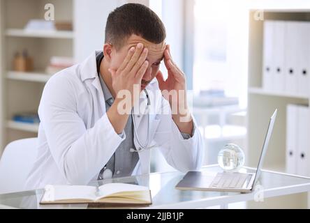 I cant think clearly with this headache. a handsome young doctor sitting alone in his clinic and feeling stressed while using his laptop. Stock Photo