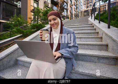 Young arab woman drinking coffee and working on a laptop outdoors Stock Photo