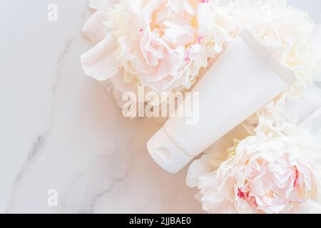 Flat lay composition with cosmetic products and peony flowers on a marble. Top view. Unbranded plastic white tube with face or hand cream or cosmetic Stock Photo