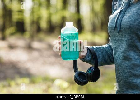 Woman drinking water from bottle and holding headphones. Resting during jogging and running in forest. Fitness and sport activity Stock Photo