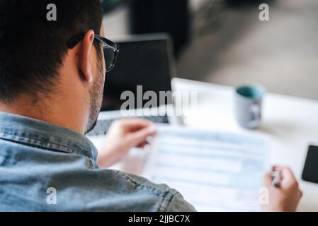 Rear view of male freelancer reading documents. Young businessman is analyzing business strategy. He is working while sitting at table in home office. Stock Photo