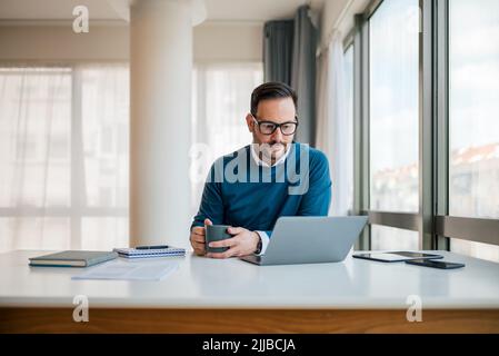 Serious thoughtful businessman analyzing plan on laptop. Male manager is having coffee while working at office desk. He is wearing smart casuals. Stock Photo