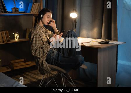 Asian teenage girl listening to the music using a cell phone and headphones at home at late night, student relaxing from study while sitting at a desk Stock Photo