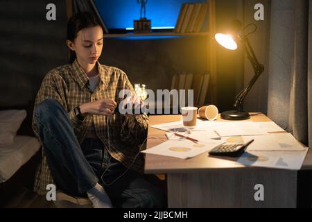 Asian female student studying at night, resting and plugging headphones into phone, relax and listening to music for relaxing at home Stock Photo