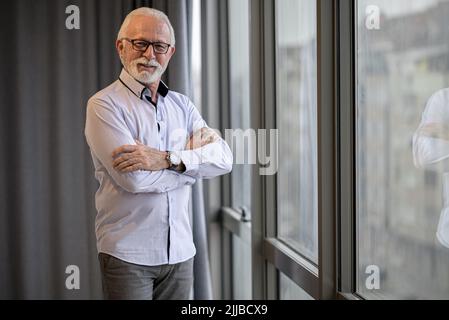 Portrait of determined senior entrepreneur by window. Confident elderly businessman is standing with arms crossed. He is wearing formals at corporate Stock Photo