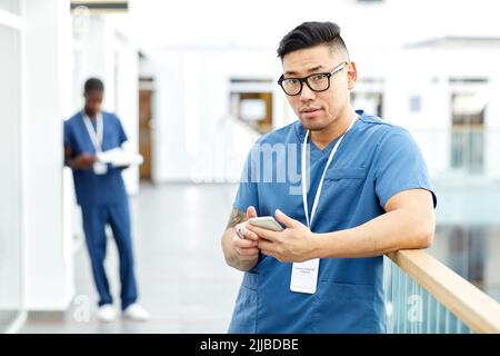 Waist up portrait of Asian male doctor looking at camera while using smartphone in hall of modern clinic, copy space Stock Photo