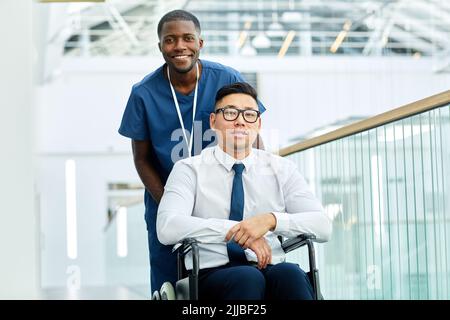 Portrait of smiling African American medic pushing wheelchair while assisting patient with disability in modern clinic, copy space Stock Photo