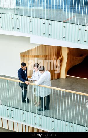 Graphic wide angle view at three doctors discussing case while standing on balcony in modern clinic, copy space Stock Photo