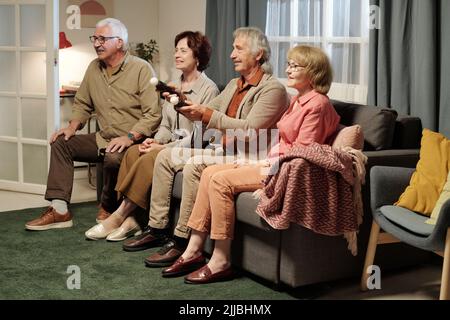 Group of four senior friends sitting on couch in front of tv set while playing leisure game with gamepads held by one of them Stock Photo