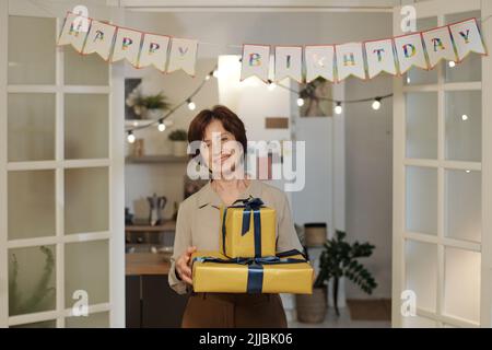 Aged cheerful woman in smart casualwear holding stack of packed presents while standing in living room during birthday party Stock Photo