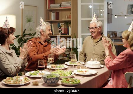 Group of cheerful senior people clapping hands while looking at their friend celebrating birthday while sitting by festive table Stock Photo