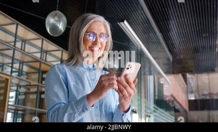 Gray-haired senior businesswoman is using a smartphone in an office. Stock Photo