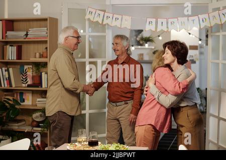Two senior women giving each other hug while their husbands shaking hands in living room with served table prepared for birthday party Stock Photo