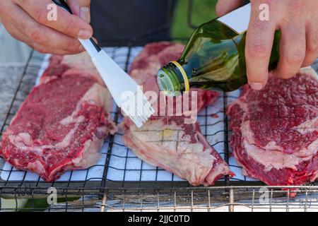 Fresh meat on barbecue metal mesh grilling basket rack. Raw beef steak, a man oil the meat. Delicious food for summer time. Stock Photo