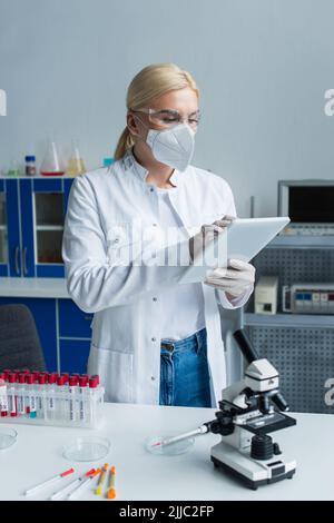 Scientist in protective mask using digital tablet near test tubes and microscope in lab Stock Photo