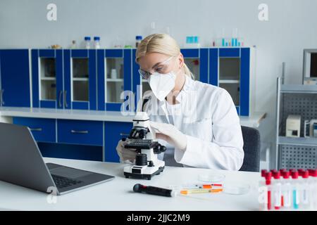 Scientist in protective mask using microscope near test tubes in laboratory Stock Photo