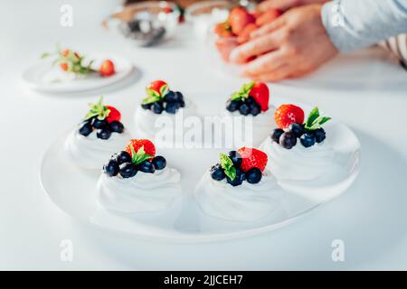 Cooking, making homemade meringue. Female hands decorating pavlova cakes with fresh berries on the kitchen table with ingredients. Recipes for delicio Stock Photo