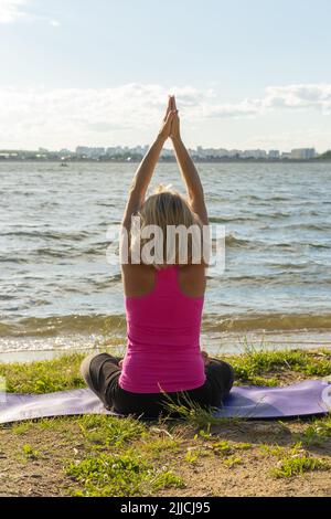 An elderly woman sitting in the lotus position and raising her hands up and looking at the lake, a view from the back. Meditation. Sports in old age. Stock Photo