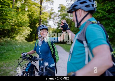 Active senior couple riding electric bicycles on road at summer park, high fiving, healthy lifestyle concept. Stock Photo