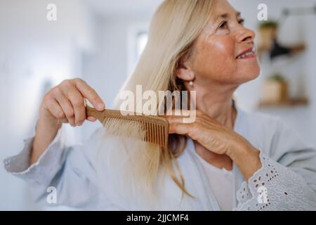 Beautiful senior woman in bathrobe combing hair with wooden comb in bathroom, sustainable lifestyle. Stock Photo