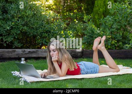 Full body view of young woman concentrated working online on red leaning on a towel in the garden grass in summer Stock Photo