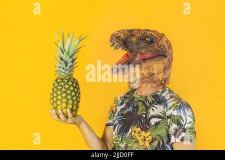 Man with dinosaur animal mask holding pineapple fruit isolated on yellow background.Copy space.Healthy food ,tropical concept. Stock Photo