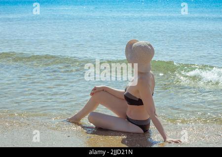 An adult slender woman in a swimsuit sits in sea water and looks at the sea. Summer vacation, rest and relaxation. Stock Photo