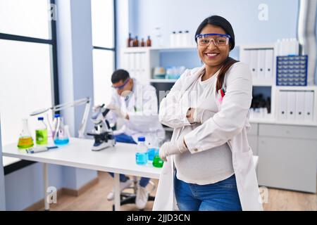 Young hispanic woman expecting a baby working at scientist laboratory happy face smiling with crossed arms looking at the camera. positive person. Stock Photo
