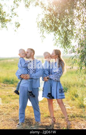 Happy parents hold their two twins sons on the hands and kiss them during walk on meadow. They are dressed in Ukrainian national embroidered shirts. Stock Photo