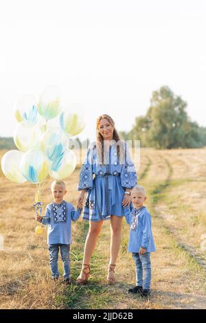 Happy mother with two twins sons walk on meadow with air balloons. They are dressed in Ukrainian national embroidered shirts. Stock Photo