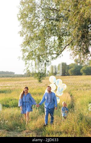 Happy parents with two their twins sons walk on meadow with air balloons in their hands. They are dressed in Ukrainian national embroidered shirts. Stock Photo