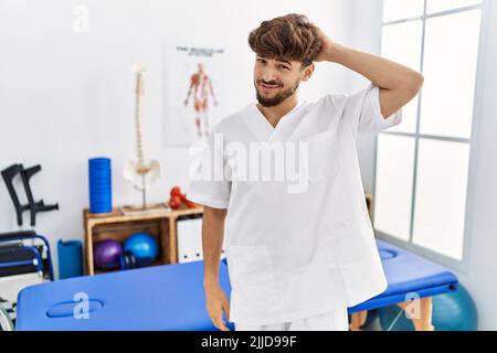 Young arab man working at pain recovery clinic confuse and wonder about question. uncertain with doubt, thinking with hand on head. pensive concept. Stock Photo