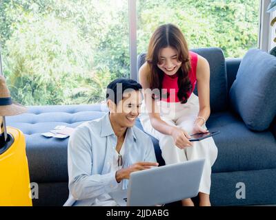 Happy Asian couple, young woman and man looking and using laptop computer and smartphone together for flight booking and trip information in living ro Stock Photo