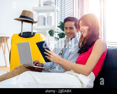 Happy Asian couple, young man and woman looking and using laptop computer and smartphone together for flight booking and trip information in living ro Stock Photo