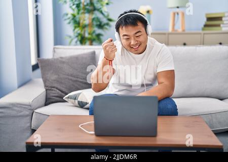 Young chinese man using laptop at home screaming proud, celebrating victory and success very excited with raised arm Stock Photo