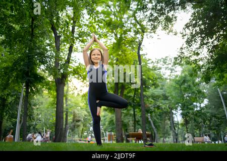 a little girl in the park is doing yoga, she stands on one leg, the other is tucked under her and stretched her arms up Stock Photo