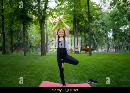 a little girl in the park is doing yoga, she stands on one leg, the other is tucked under her and stretched her arms up Stock Photo