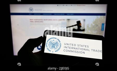 Person holding cellphone with logo of American International Trade Commission (USITC) on screen in front of webpage. Focus on phone display. Stock Photo