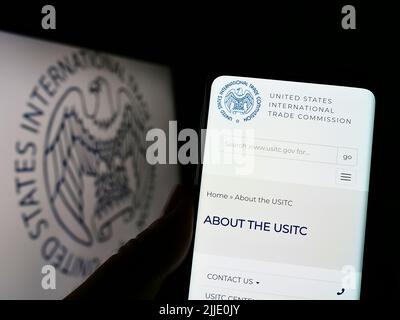 Person holding cellphone with website of US International Trade Commission (USITC) on screen in front of logo. Focus on center of phone display. Stock Photo