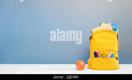 Backpack with different colorful stationery and apple on table and blue background. Banner design back to school concept background. Stock Photo