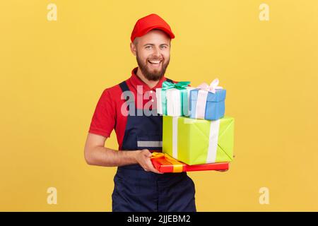 Portrait of positive happy delivery man wearing uniform and cap holding many present boxes, bringing order for celebrating holiday. Indoor studio shot isolated on yellow background Stock Photo