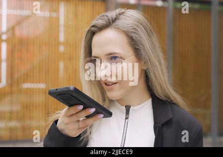 Happy girl records an audio message on a smartphone. Calls on the speaker, gives a command to a virtual assistant, uses a voice recognition applicatio Stock Photo