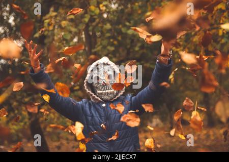 Defocus Halloween people. Person in grim reaper mask raising hand and throwing leaves. Many flying orange, yellow, green dry leaves. Skull ghost. Fall Stock Photo