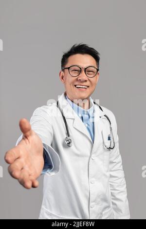 Smiling mature asian man doctor in white coat, glasses with stethoscope give his hand, greets patient in camera Stock Photo