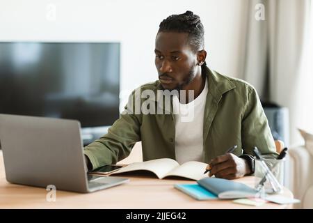 Focused african american man using laptop, working remotely and taking notes, having remote meeting from home Stock Photo