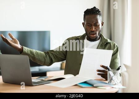 Shocked black man reading paper letter mail and spreading hands, sitting at table with laptop at home office Stock Photo
