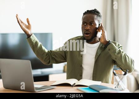 Discontented black man talking on cellphone and using pc, having problem with internet connection at home Stock Photo