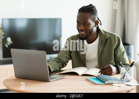Excited black guy looking for job online, sitting at table, using laptop computer and taking notes, copy space Stock Photo