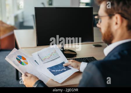 Successful entrepreneur reading financial reports, sitting at workplace with computer with black empty screen Stock Photo